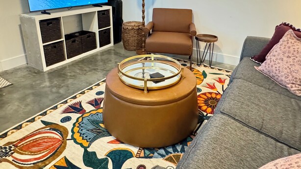 Una sala de estar con televisor, un sillón marrón, un sofá, un tapete estampado y una mesita auxiliar.