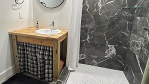 Un baño con tocador de madera, un espejo redondo, ducha con puerta de cristal y piso estampado.
