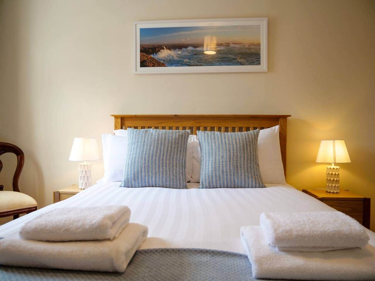 A hotel room with a bed, pillows, a nightstand with a lamp, a chair, and a framed picture on the wall.