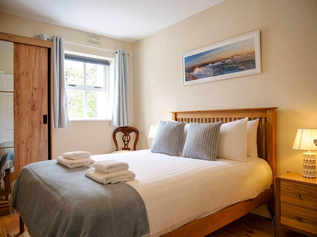 Une chambre à coucher avec un lit en bois, une table de chevet, une chaise, une fenêtre avec des rideaux et un tableau accroché au mur.
