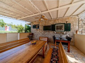A covered outdoor seating area with stone walls, wooden benches, a table, and a ceiling fan. - Natur Olive House Ston (Ston)