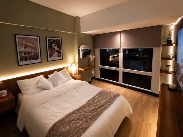 Hotelkamer met een groot bed, nachtkastje, lamp, raam met uitzicht en foto’s in lijsten aan de muur.