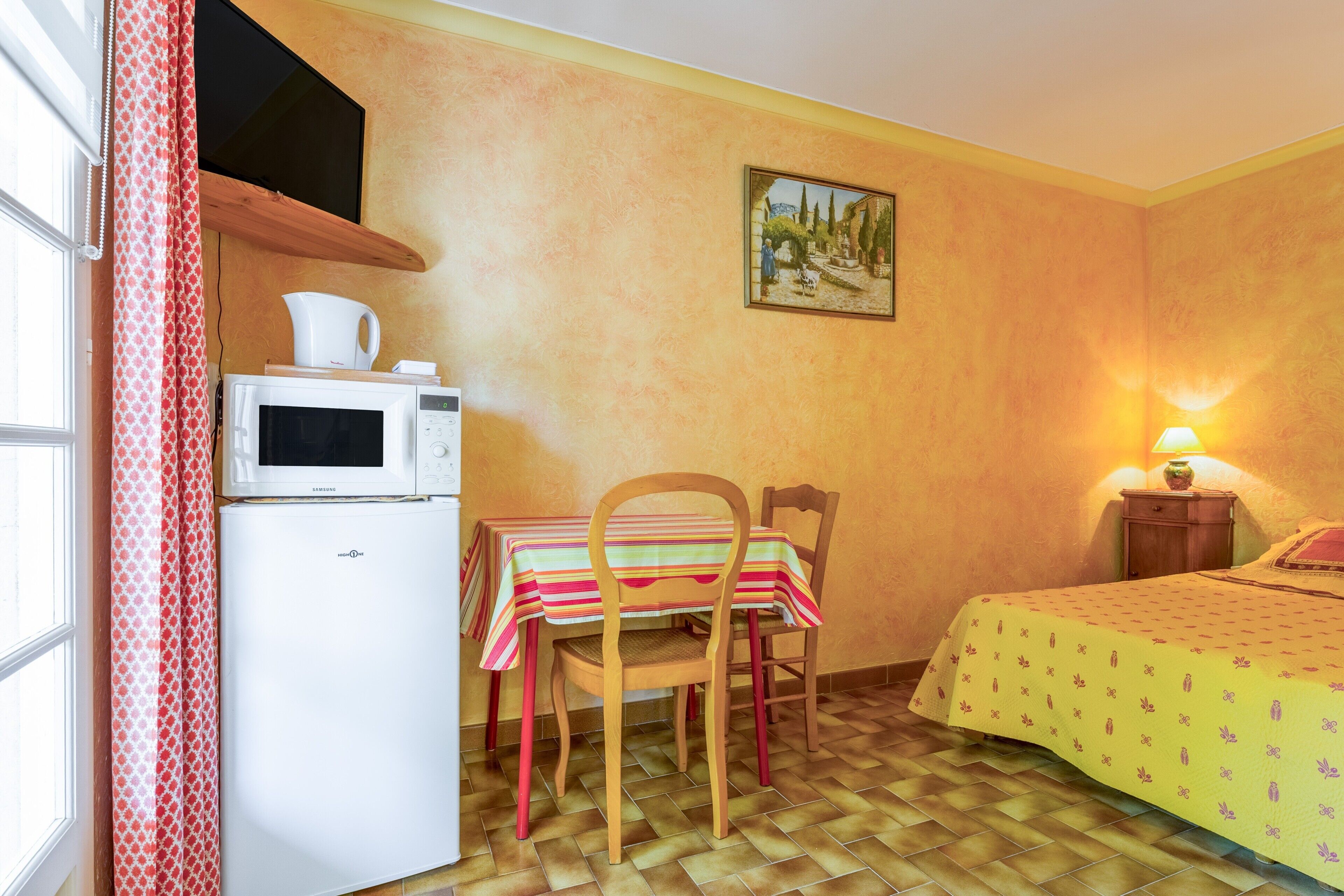 Une pièce compacte comprenant un coin cuisine, une table à manger avec des chaises, un lit avec une couvre-lit à motifs, un tableau au mur et une fenêtre avec des rideaux.