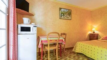 Une pièce compacte comprenant un coin cuisine, une table à manger avec des chaises, un lit avec une couvre-lit à motifs, un tableau au mur et une fenêtre avec des rideaux.