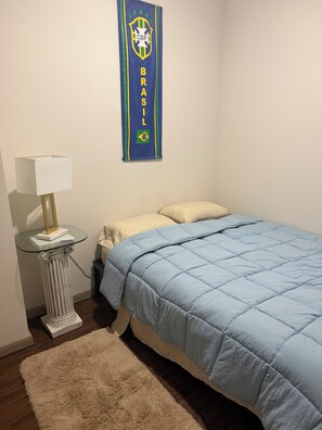 Een slaapkamer met een bed, nachtkastje, lamp, wanddecoratie en vloerkleed.