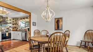 Een eetgedeelte met een houten tafel en stoelen, een kroonluchter, en een keuken op de achtergrond.