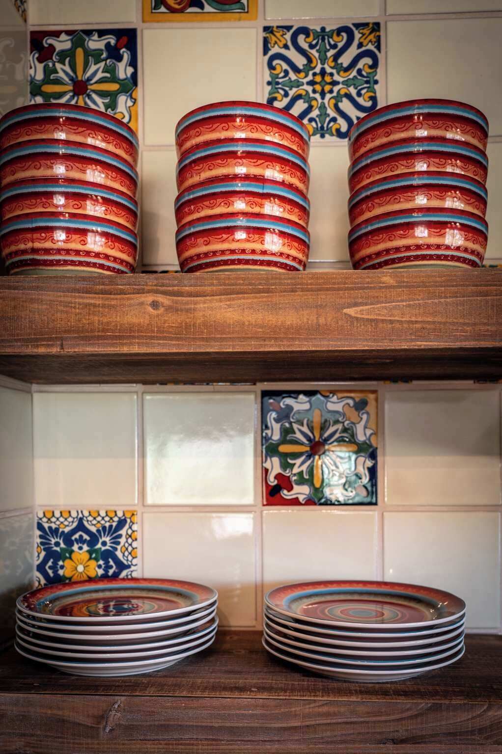 Une cuisine avec des étagères en bois où sont empilées des assiettes aux couleurs vives, et des carreaux décoratifs ornant le mur.