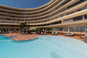 Ein mehrstöckiges Hotel mit einem zentral gelegenen Schwimmbecken, umgeben von Liegestühlen und Sonnenschirmen.