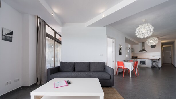 Una sala de estar moderna con un sofá gris, una mesita de centro blanca y un comedor con sillas rojas.