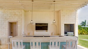 An outdoor kitchen area with a large island, bar stools, a flat-screen TV, and a grill. - Luxury 8BR Villa Hacienda  Punta Cana (Punta Cana)