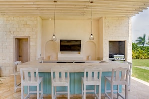 Un área de cocina al aire libre con una isla grande, taburetes de bar, un televisor de pantalla plana y una parrilla.