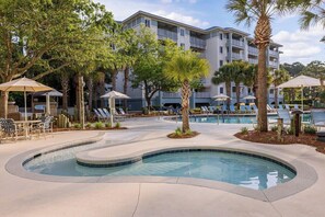 A pool area with lounge chairs and umbrellas, surrounded by trees and a building with balconies. - 2 Bedroom Villa Beach Access at SurfWatch Resort (Hilton Head Island)
