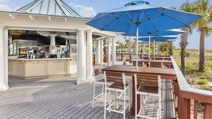 Ein überdachter Außenbereich mit Holztischen und -stühlen, eine Bar mit der Beschriftung 'SAI SALTY' und im Hintergrund Palmen.
