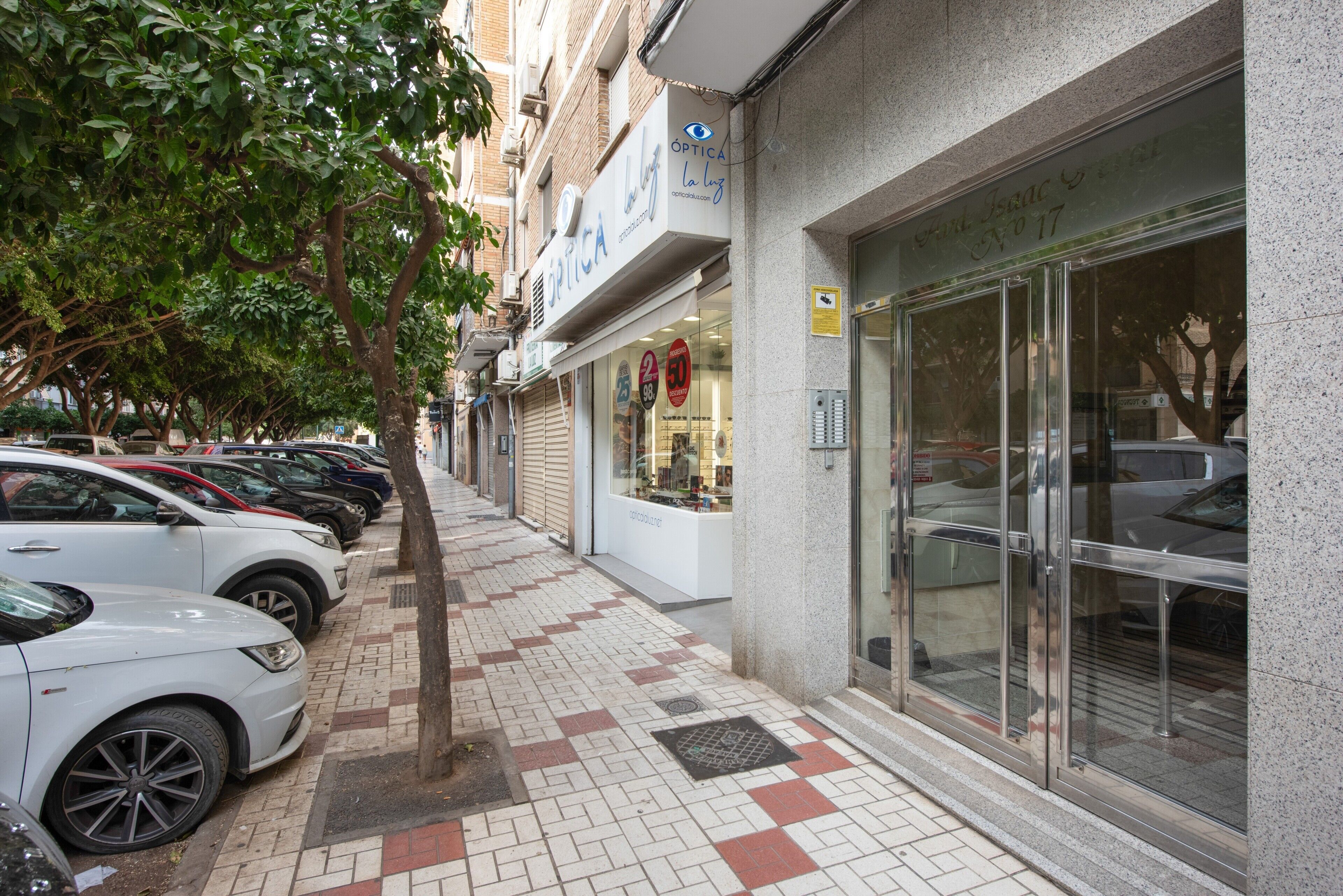 Una acera con autos estacionados, una fachada comercial con un letrero y un árbol.