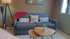 Ein Zimmer mit grauem Sofa, zwei gemusterten Kissen, einer roten Decke, einem kleinen runden Holztisch mit einer Pflanze und einer Stehlampe.