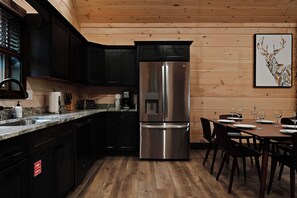 A modern kitchen with dark cabinets, a stainless steel refrigerator, a granite countertop, and a dining area with a wooden table and chairs. - NEW CABIN: Sauna | Hot Tub | Fire Pit | EV (Gatlinburg)
