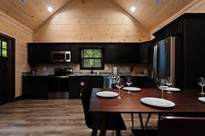 A modern kitchen with dark cabinetry, stainless steel appliances, and a dining area with a wooden table set for four. - NEW CABIN: Sauna | Hot Tub | Fire Pit | EV (Gatlinburg)