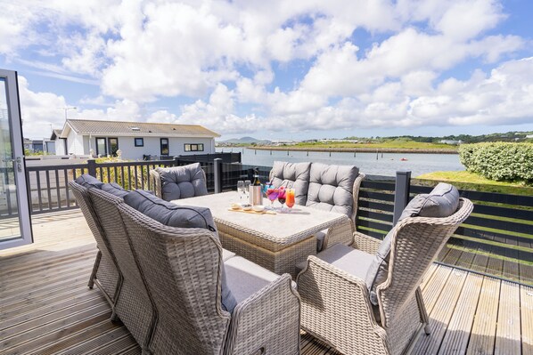 Une terrasse dotée de meubles en osier et d’une table dressée pour le repas, offrant une vue sur une étendue d’eau et un paysage lointain.
