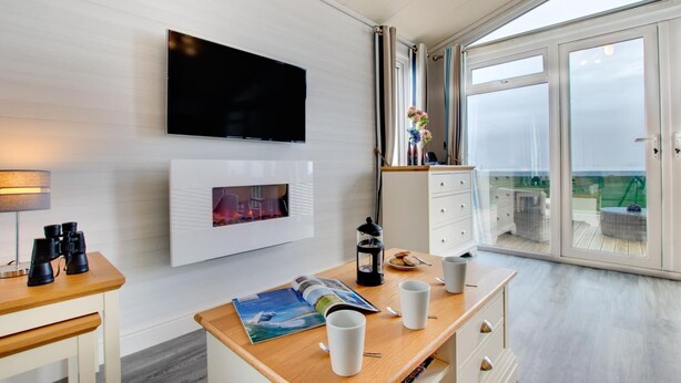 Quarto moderno com televisão de tela plana, lareira, área de jantar com mesa e cadeiras, e uma grande porta de vidro deslizante que dá para uma área externa.