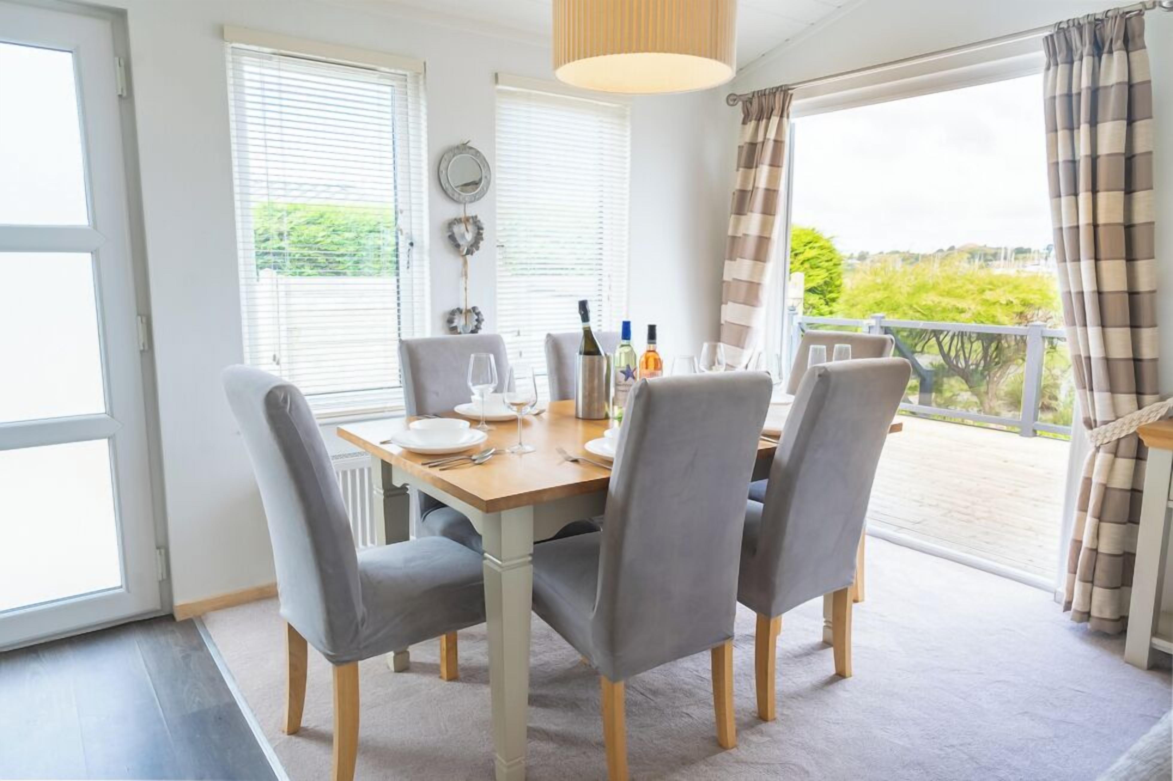 Un comedor con una mesa dispuesta para cuatro personas, sillas, una ventana con persianas y vistas al exterior con vegetación.