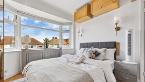 A bedroom with a large window, a bed with multiple pillows, a bedside table, a wall-mounted lamp, and a radiator. - Cozy 3bd, Garden, Free Parking, M25,bluewater (Dartford)