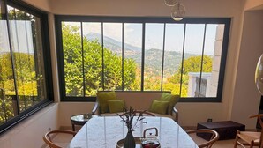 A dining area with a marble table, wooden chairs, and a view of greenery and mountains through large windows. - Villa Apolline · 1892 Estate · 5 BR, Heated Pool, Riviera Views (Grasse)