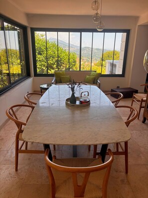 Ein Essbereich mit einem Marmortisch, Holzsesseln und Blick auf Grünflächen und Berge durch große Fenster.