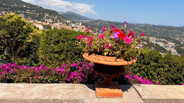 A terracotta pot with flowers, a stone wall, and a view of a town with buildings and hills in the background. - Villa Apolline · 1892 Estate · 5 BR, Heated Pool, Riviera Views (Grasse)
