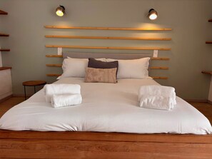 Ein ordentlich bezogenes Bett mit weißen Leinen und zwei weißen Handtüchern, ein Kopfteil mit horizontalen Holzleisten sowie zwei Wandleuchten.