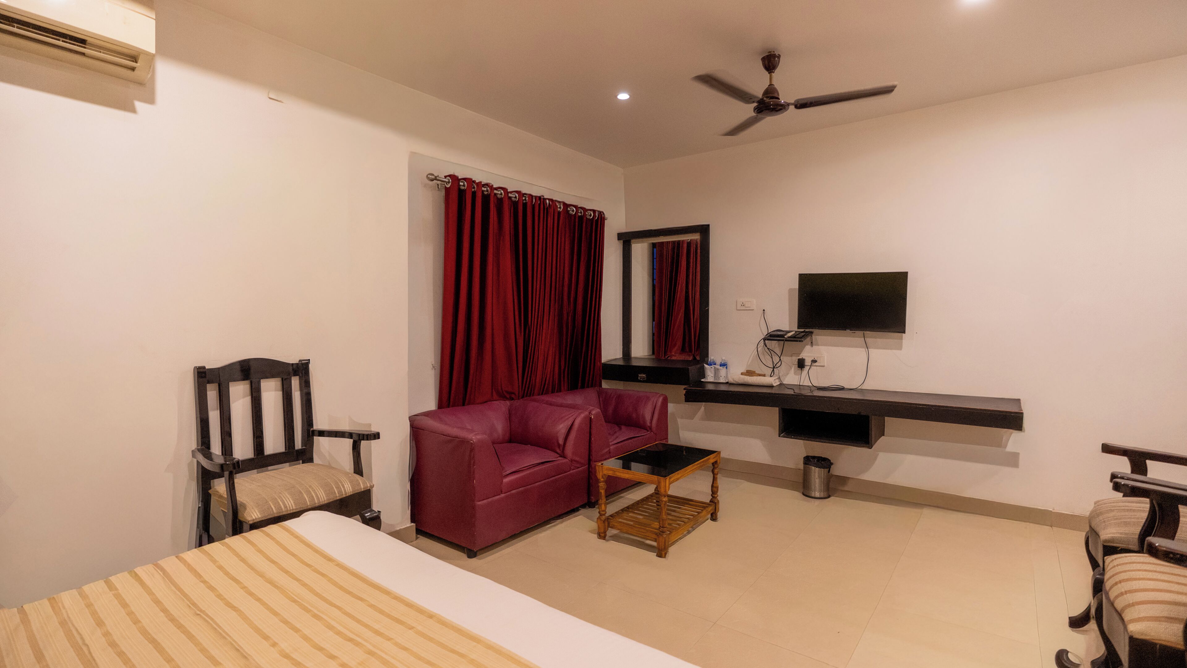 A hotel room with a bed, a red sofa, a wooden coffee table, a TV mounted on the wall, and a ceiling fan.