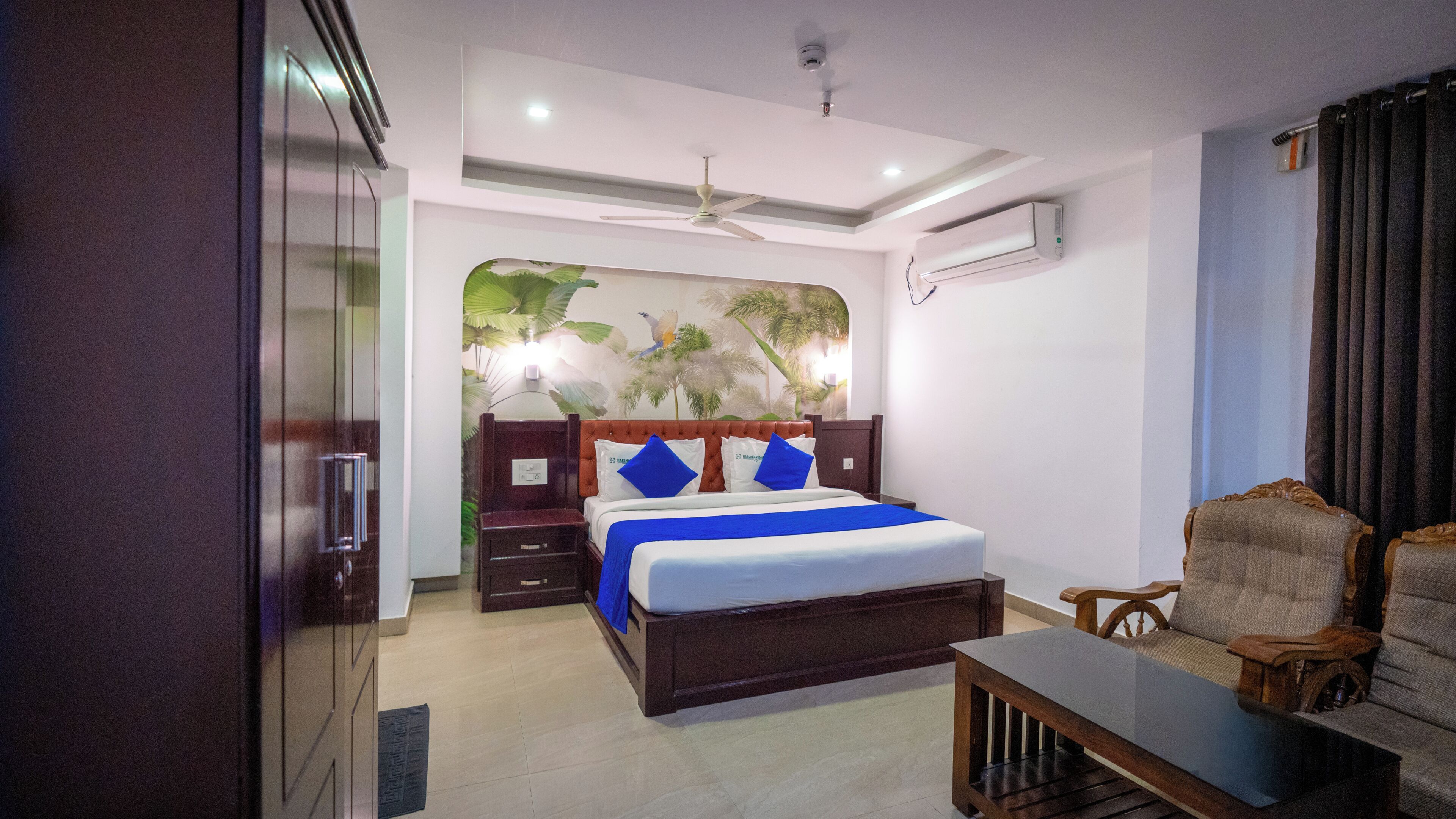 A hotel room with a bed, a wooden nightstand, a desk, a chair, a ceiling fan, and a wall-mounted air conditioner.