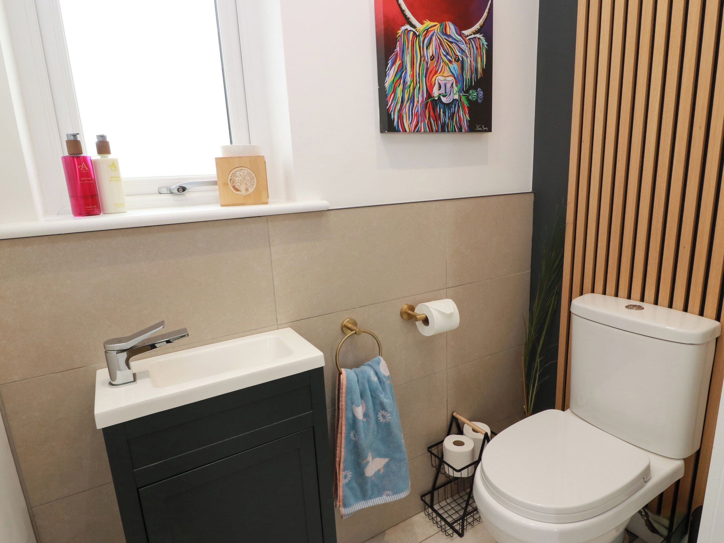 A bathroom with a white sink, a toilet, a window with a view, and a shelf with toiletries.