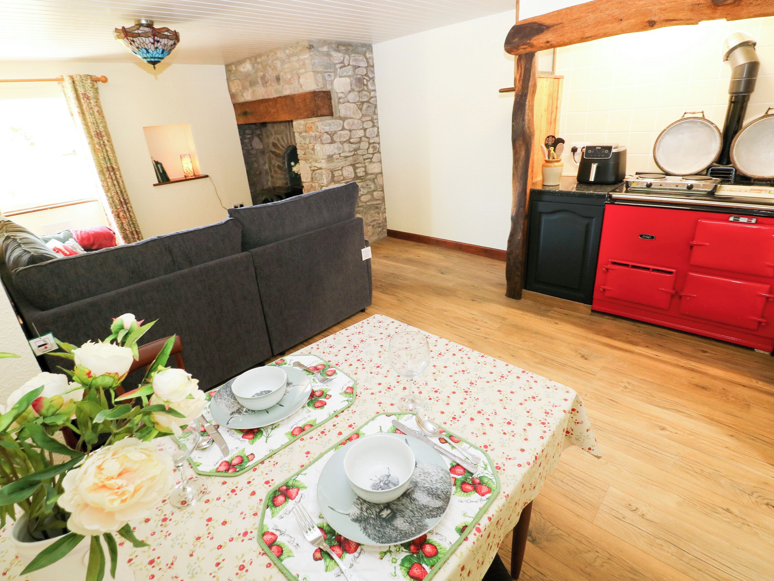 A dining area with a table set for two, a sofa, a stone fireplace, and a red kitchen with a stove.