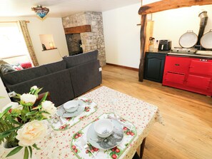 A dining area with a table set for two, a sofa, a stone fireplace, and a red kitchen with a stove. - Dolcoed (Llandeilo)