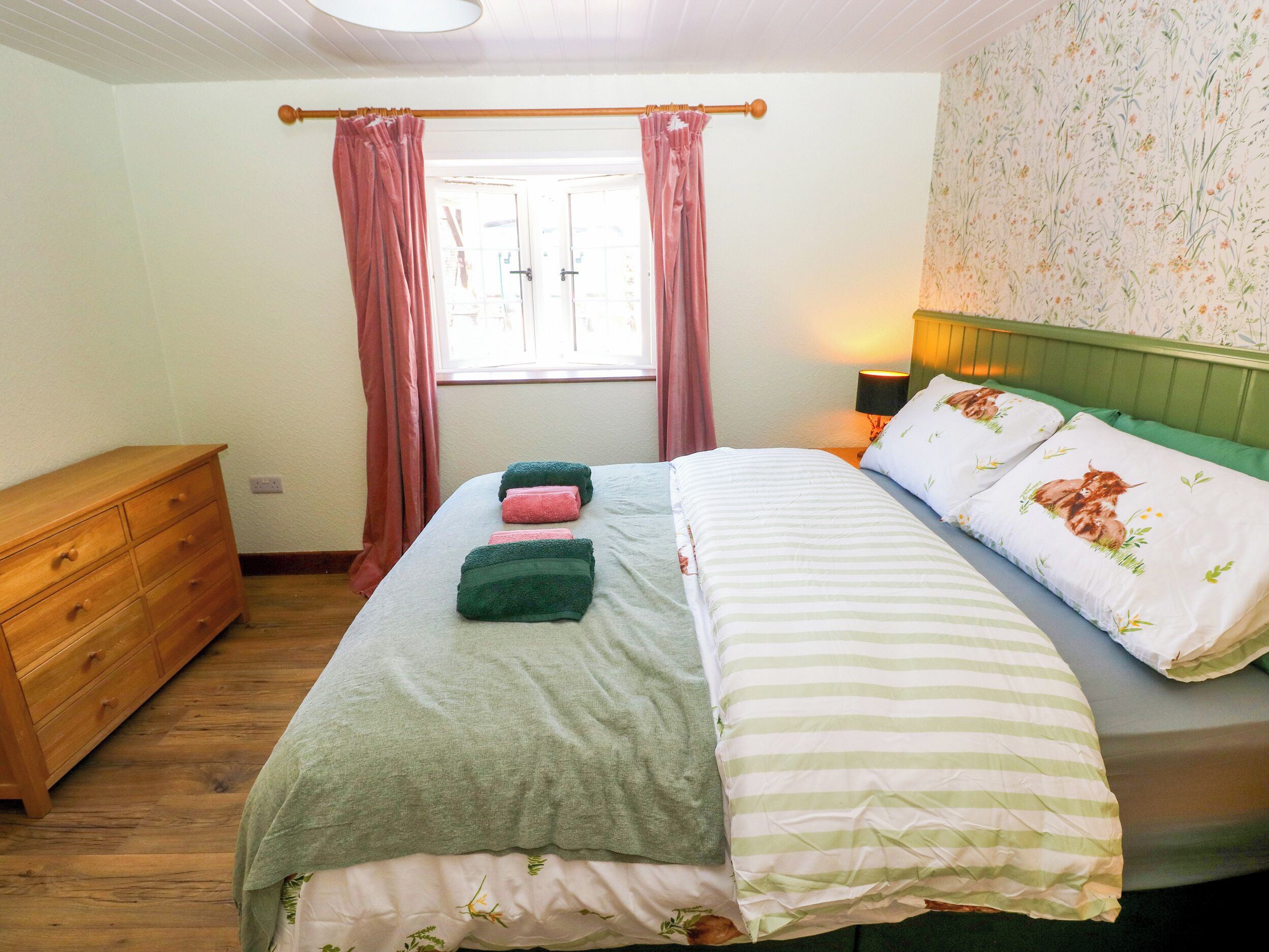 Ein Schlafzimmer mit einem Bett, einer hölzernen Kommode, einem Fenster mit Vorhängen und einer Wand mit Blumenmuster-Tapete.