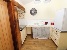A compact kitchen with wooden cabinets, a white countertop, a red kettle, and a clock on the wall.