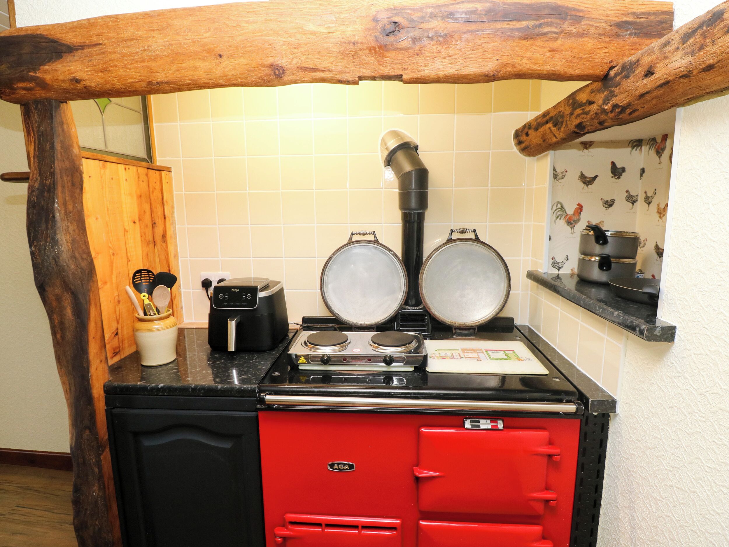 Una cocina con una estufa AGA roja, dos sartenes, una tetera eléctrica negra y un armario de madera.