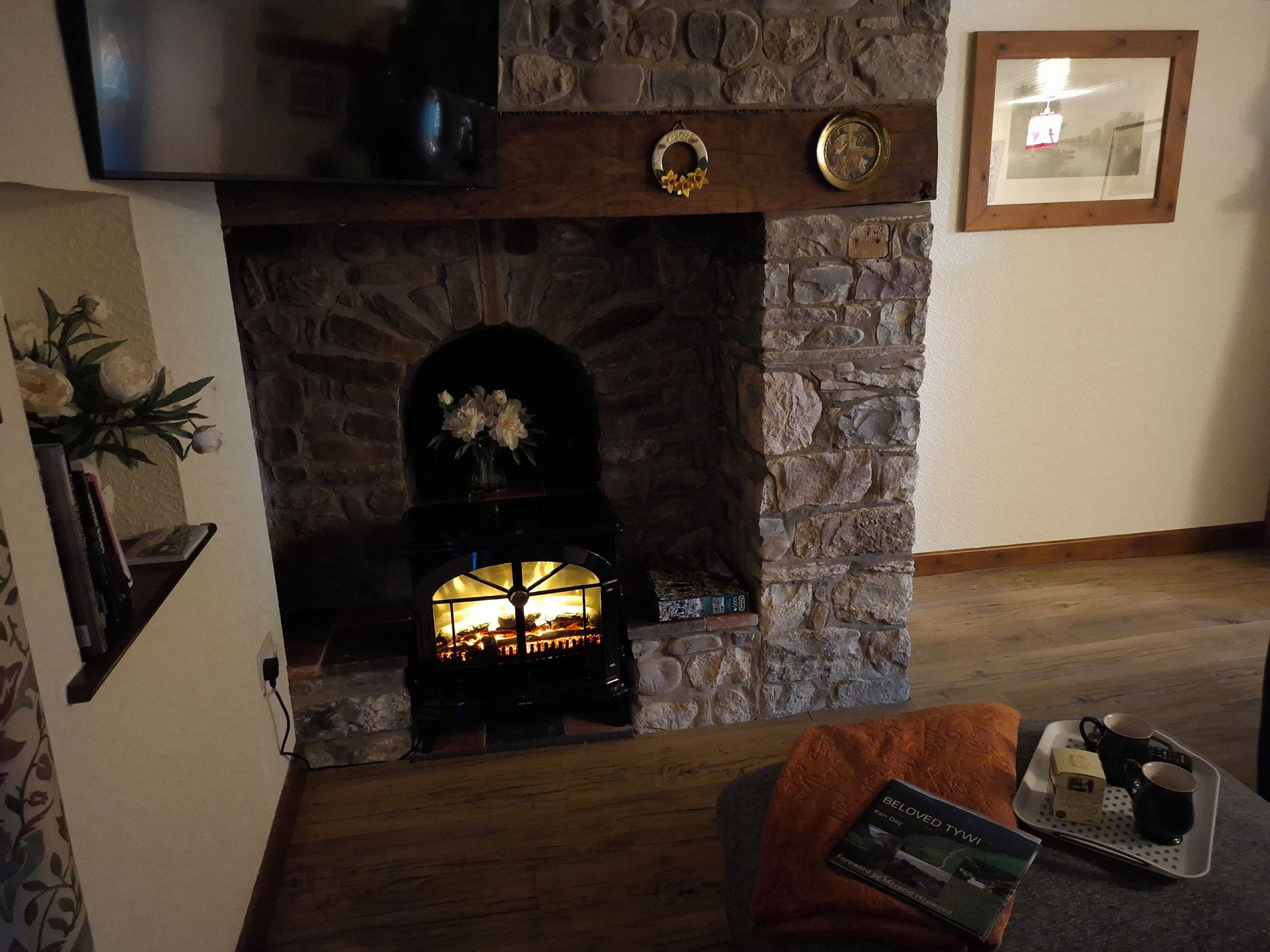 A stone fireplace with a fire, a framed picture on the wall, and a book on a tray.