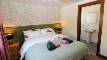 Une chambre à coucher avec un lit, deux lampes de chevet, une tête de lit verte, un papier peint à motifs floraux, une porte en bois et une salle de bain avec un lavabo et un miroir.