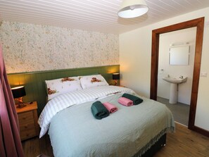A bedroom with a bed, two bedside lamps, a green headboard, a floral wallpaper, a wooden door, and a bathroom with a sink and mirror. - Dolcoed (Llandeilo)