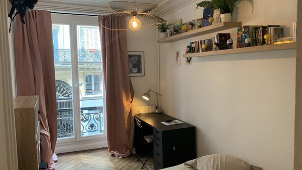 Een kamer met een bureau, stoel, boekenkast, potplant en een raam met gordijnen.