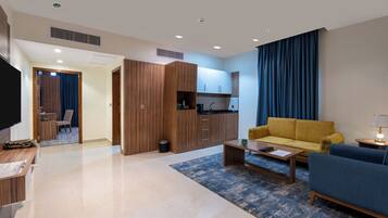 A modern living room with a sofa, coffee table, and a TV. There is a kitchen area with cabinets and a sink. The room has a large window with curtains.