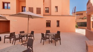 An outdoor seating area with tables and chairs, a covered umbrella, and a building with multiple windows and balconies.