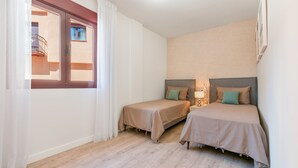 A hotel room with two beds, a window with curtains, a wooden floor, and a wall-mounted lamp.