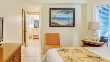 A hotel room with a bed, a chair, a desk with a lamp, a painting of a beach scene, and a television.