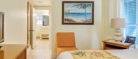A hotel room with a bed, a chair, a desk with a lamp, a painting of a beach scene, and a television.