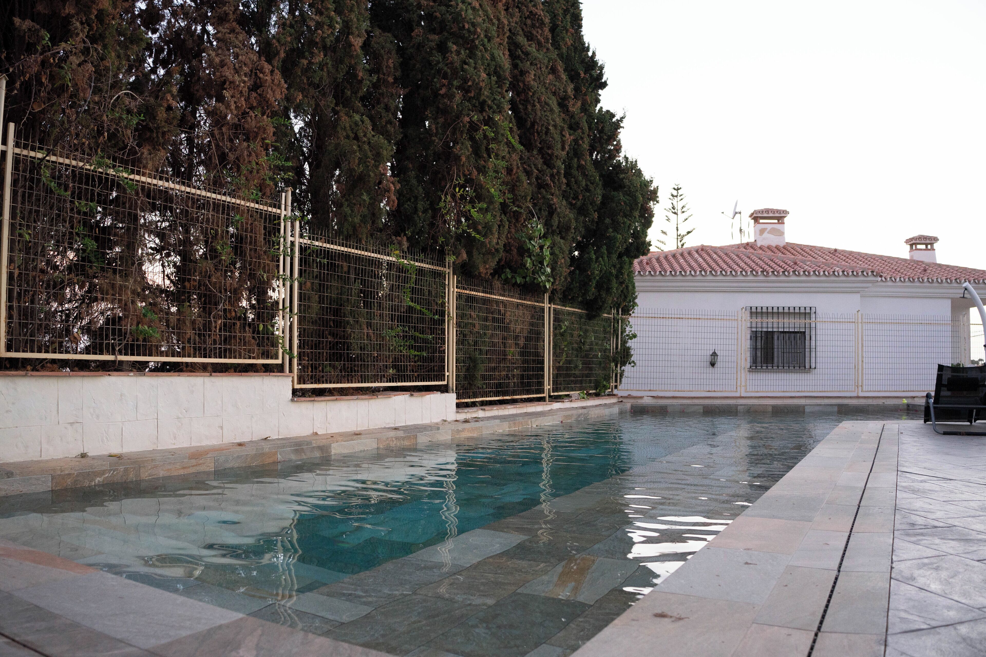 Una piscina con borde de azulejos, una zona vallada y un edificio con tejado de cerámica al fondo.