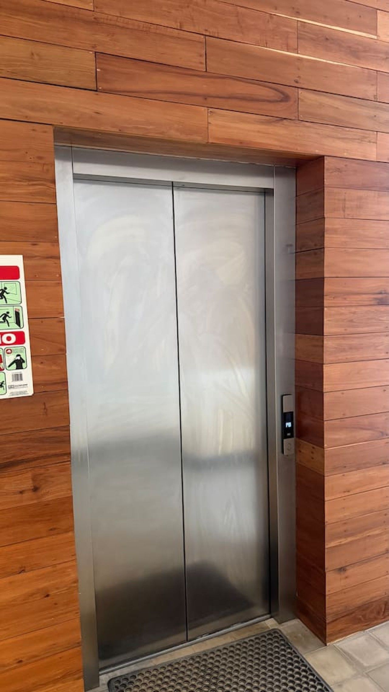 Une porte d’ascenseur métallique, dotée d’un panneau de commande sur le côté droit, est encadrée par un mur en bois sur lequel est apposé un panneau de signalisation de sécurité sur le côté gauche.