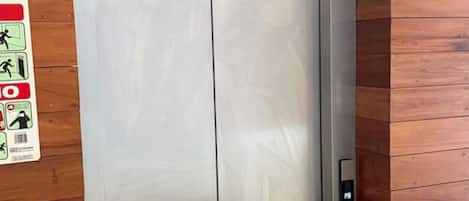 A metallic elevator door with a control panel on the right side, set against a wooden wall with a safety sign on the left.