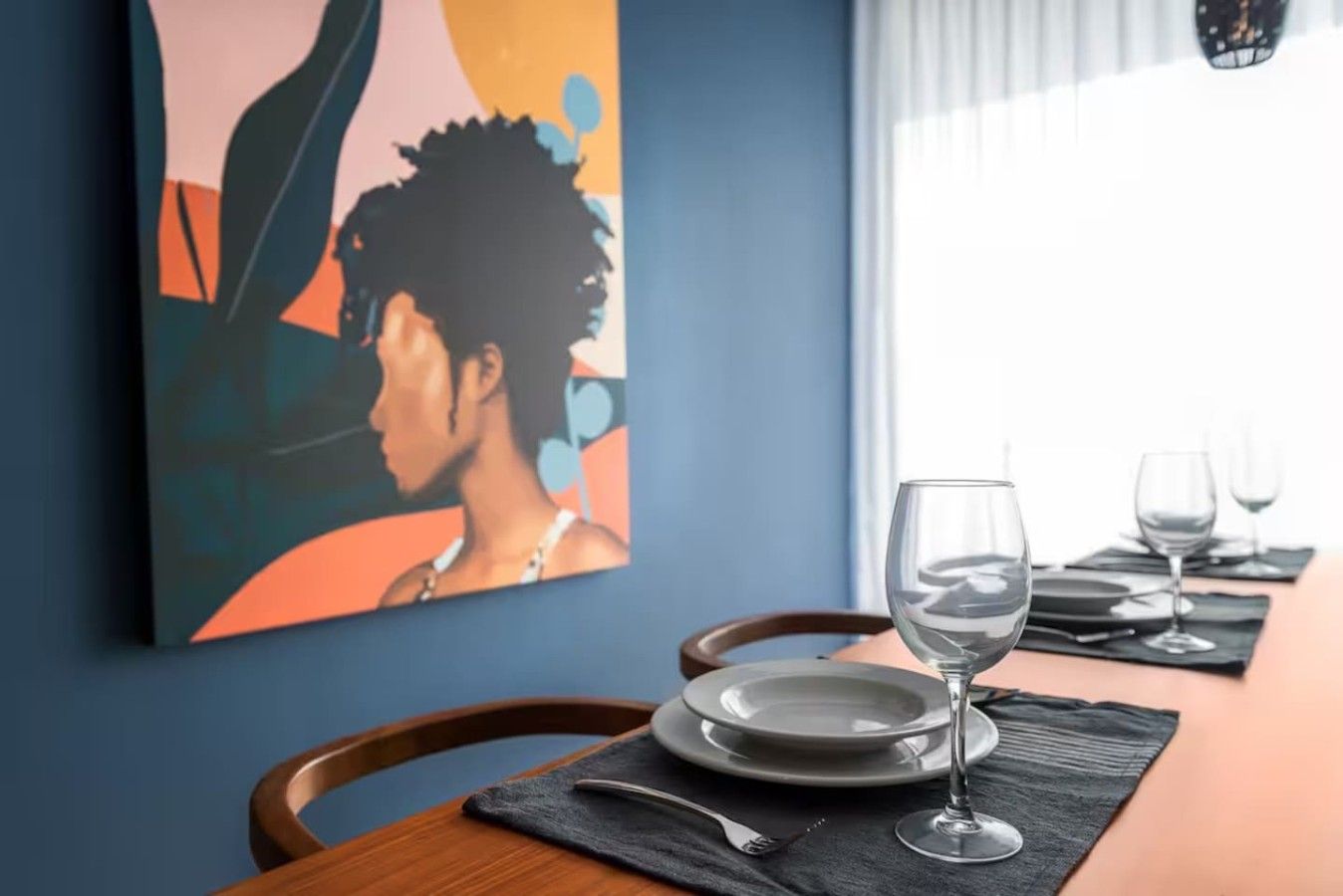 Une table à manger est dressée avec des assiettes, des couverts et des verres à vin, prêts pour quatre personnes. Un tableau coloré est accroché au mur derrière la table.
